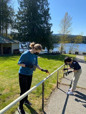 Two individuals painting a metal railing outdoors near a lake, with green grass, trees, and water in the background.