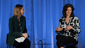 Two people talking on stage with blue curtain backdrop.