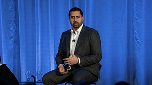 Person in suit speaking on stage with blue curtain backdrop.