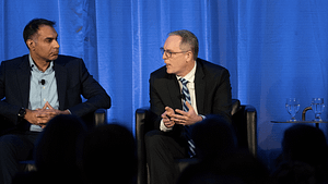 Two people seated on stage with blue curtain backdrop.