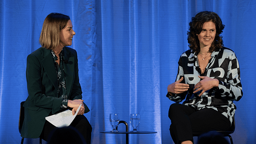 Two people talking on stage with blue curtain backdrop.