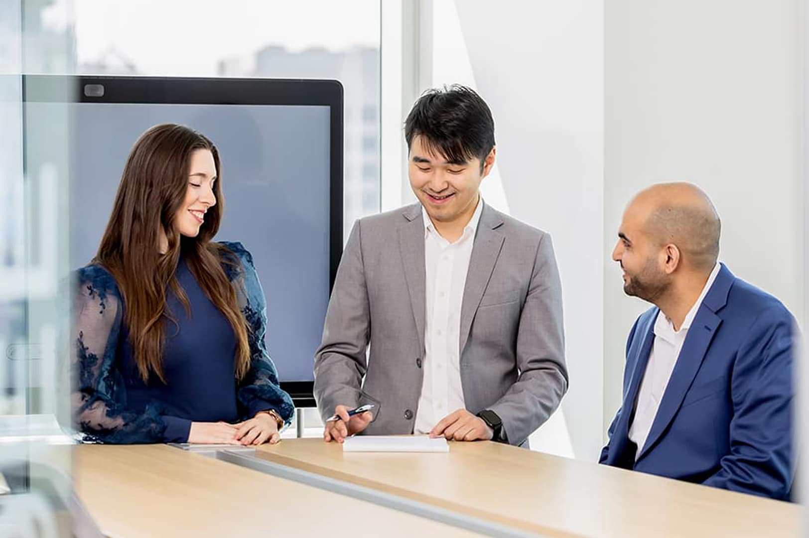 Three people standing at a conference table in an office building, smiling and collaborating Three people standing at a conference table in an office building, smiling and collaborating
