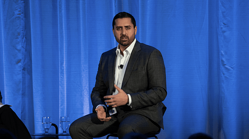 Person in suit speaking on stage with blue curtain backdrop.