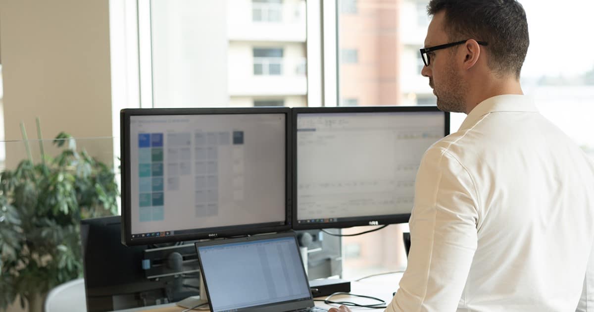 Man with glasses and light coloured shirt in working at a stand up desk with multiple monitors